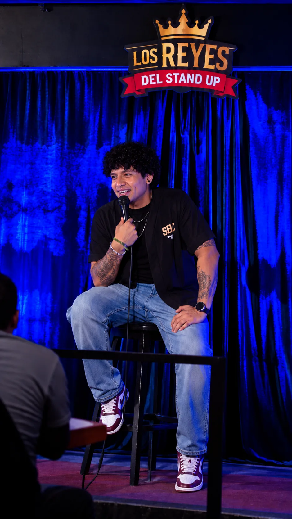 Juan sentado en un banco con micrófono en el escenario de Los Reyes del Stand Up